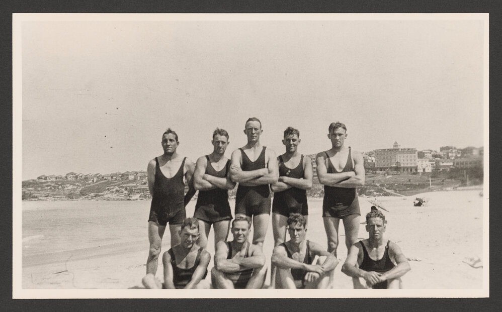 Bathers, group of men in bathing costumes, 5 standing, 4 sitting. At Bondi beach. Hotel Astra in background on right.