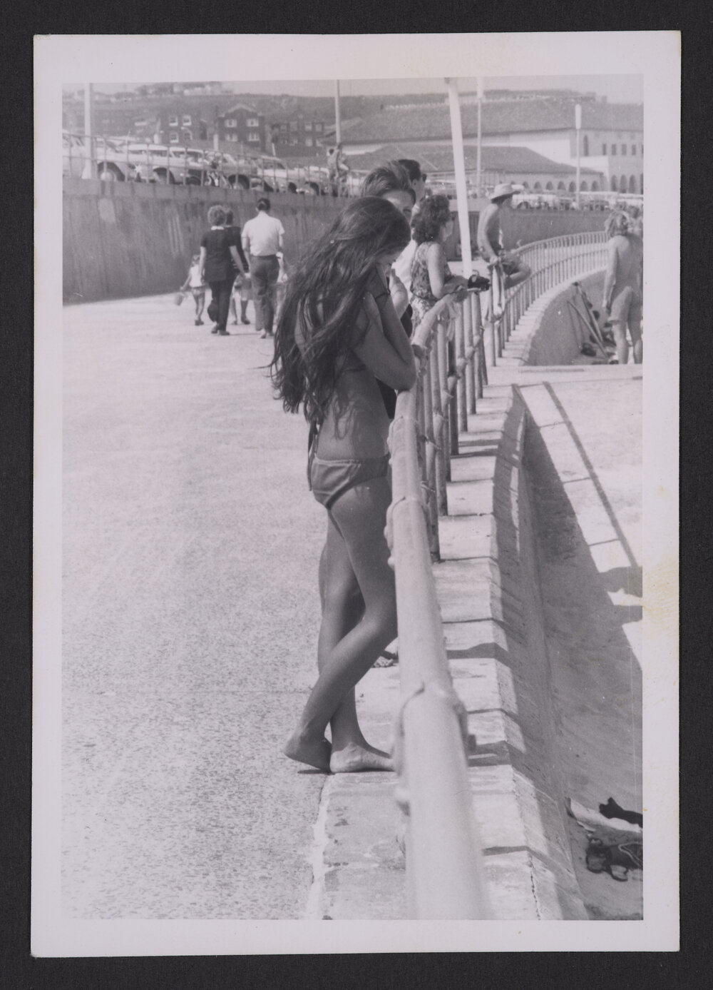 Bathers, girl in Bikini leaning on Promenade railing, Bondi beach.
