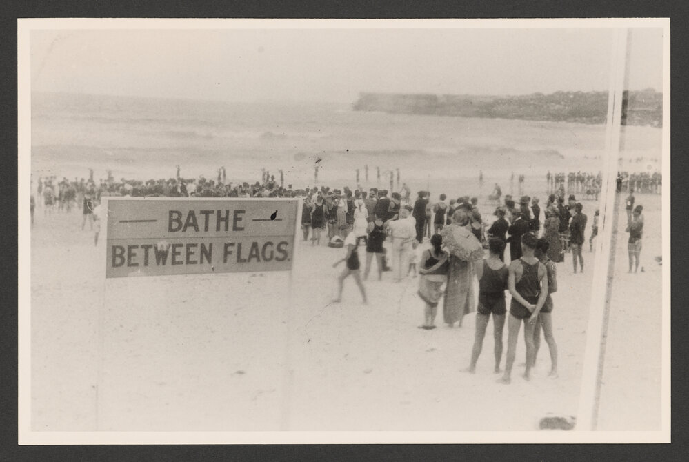 Bathers on Bondi beach watching, "Bathe between the flags" sign.