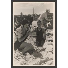 Bathers on Bondi beach. Women and boy seated on sand. Deckchairs behind them.