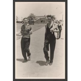 Bathers, two men fully dressed and carrying towels, head for Bondi beach. Marine drive bridge in background.