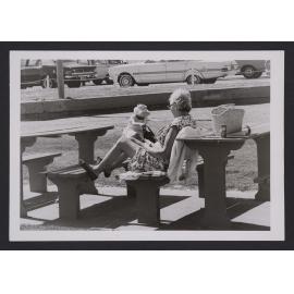 Bathers, woman seated at Picnic table and bench in Bondi beach park.
